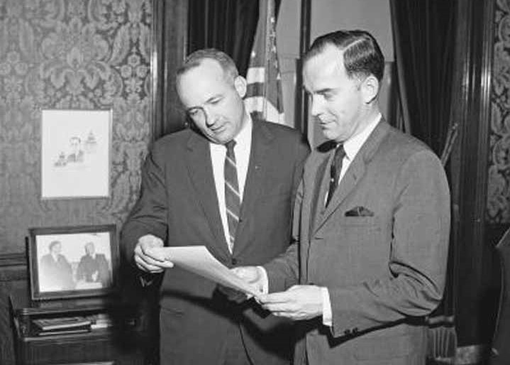 Two men in mid-1960s suits and ties hold and look at a piece of paper in a fancy office.