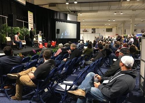 Crowd of conference attendees and three presenters watch video playing on large screen.