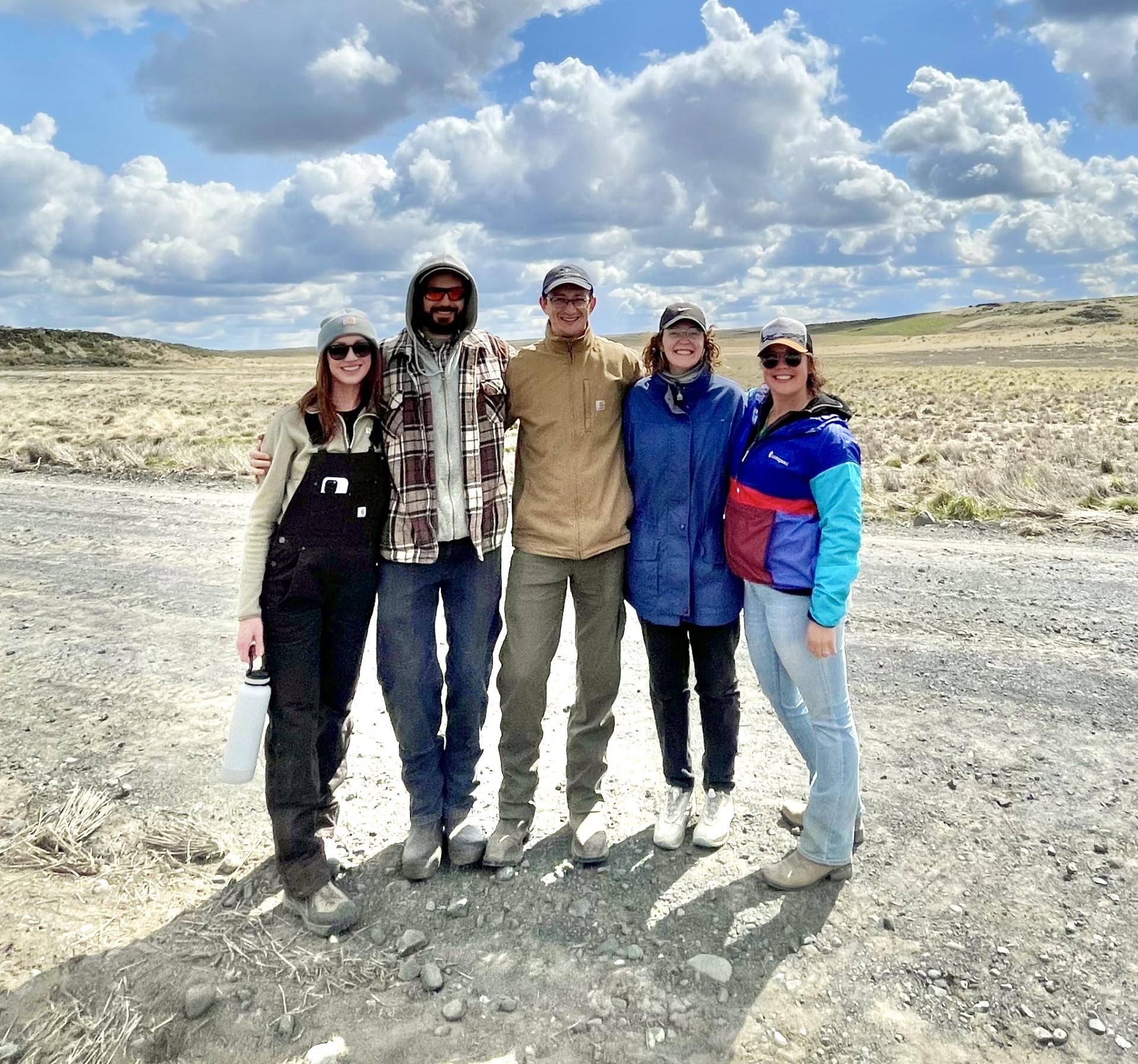 Five people with arms around each other smile at the camera. They stand on a gray gravel road in front of a field and brown hills.