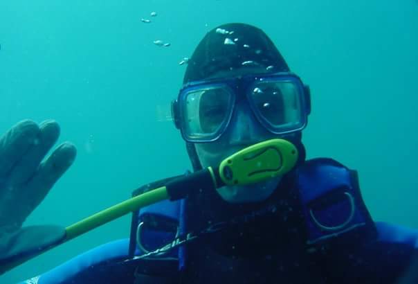 A SCUBA diver in wetsuit and hood waves at the camera.