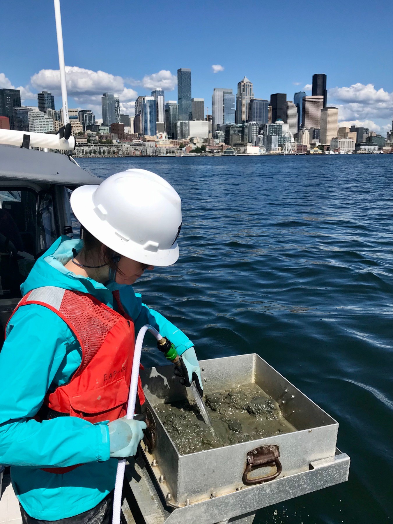 A female scientist in hard hat and life jacket works on the back deck of a boat in front of a city skyline