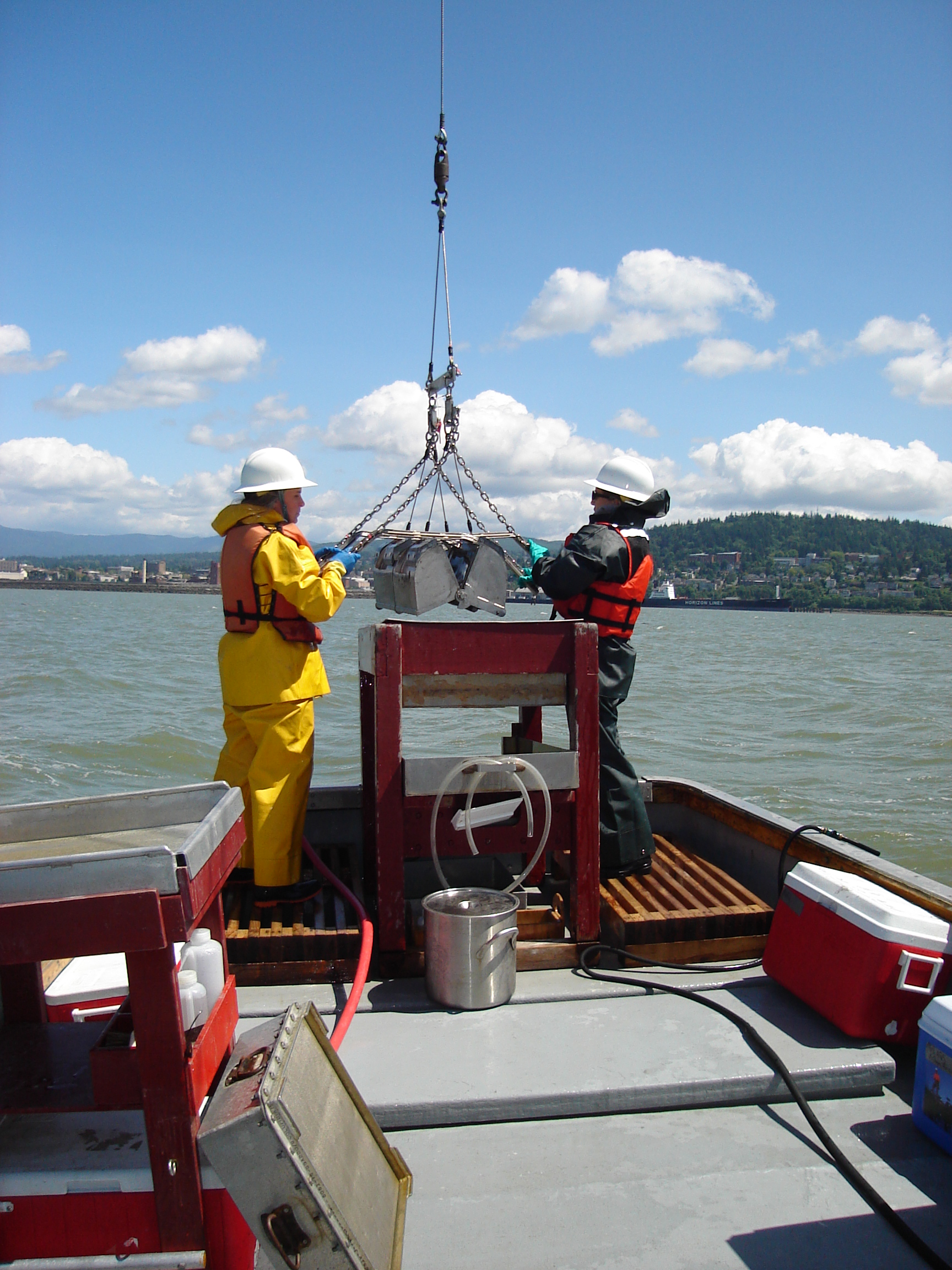 Two scientists stand at the stern of a research vessel controlling a grab sampler, like a double-scoop construction shovel, but smaller.