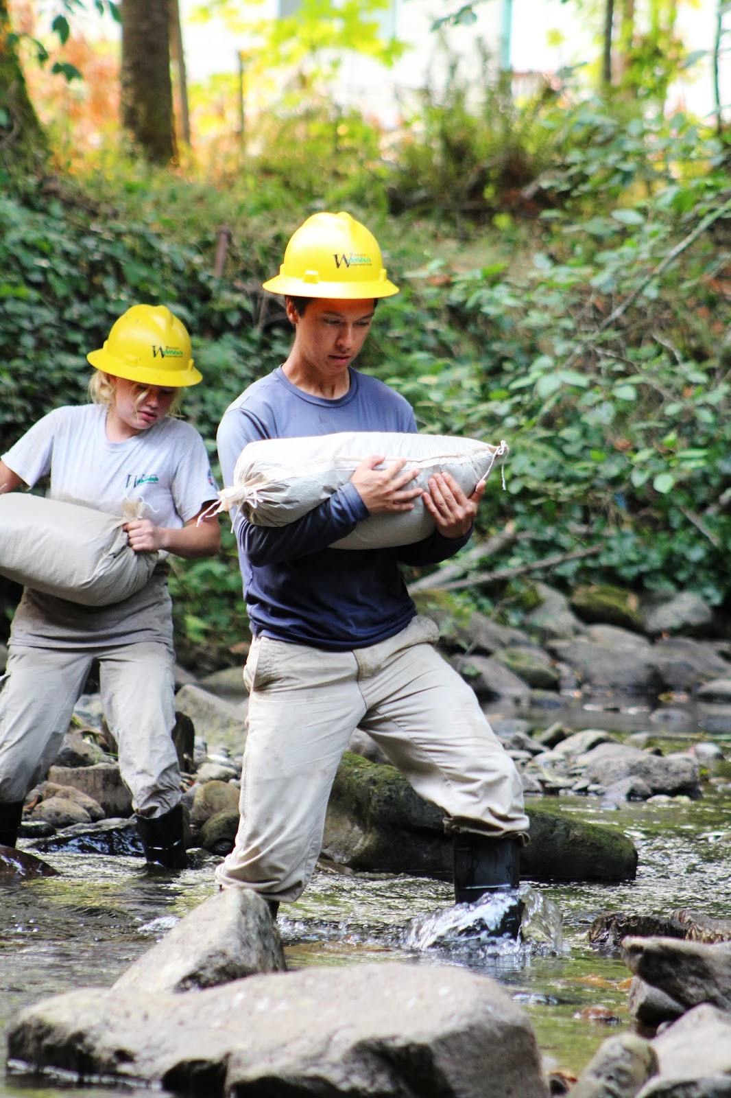 Two AmeriCorps members wearing yellow hard hats carry sand bags through a rocky river bed. 