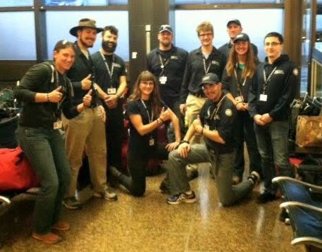 WCC AmeriCorps members gather for a group photo in an airport. They are wearing blue shirts and many are giving a thumbs up.