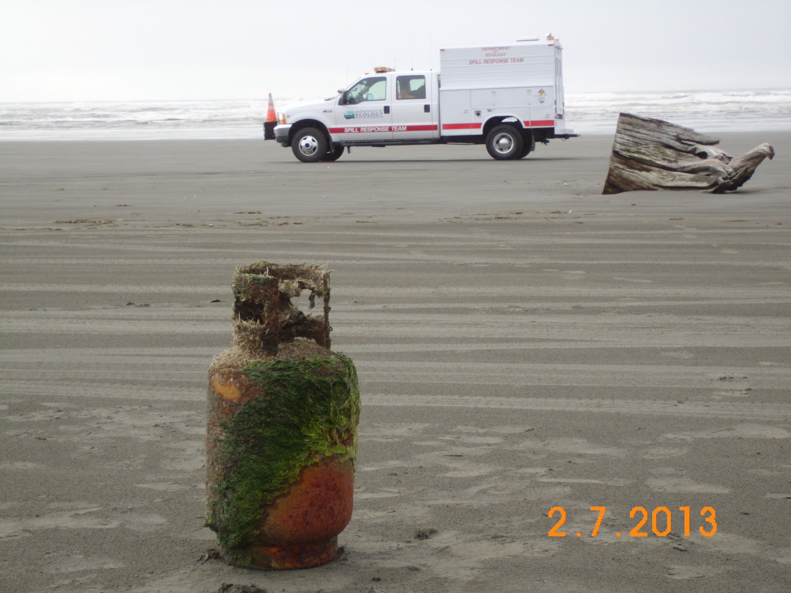 Closeup of sand and moss covered propane tank on the beach with Ecology Spills vehicle in the distance