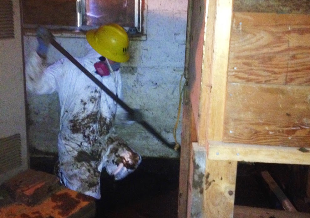 An AmeriCorps member wearing a white Tyvek suit and yellow hard hat sweeps inside a mucked and gutted home.