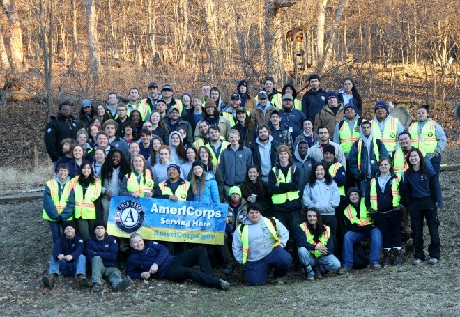 A large group of over 50 AmeriCorps members gather for a group photo near a wooded area. They hold an AmeriCorps banner in front.