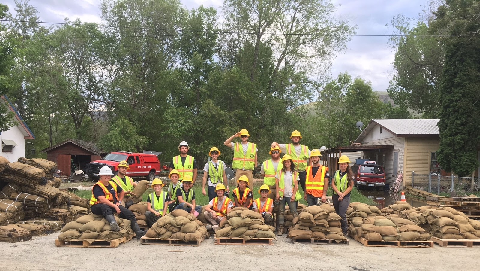 Over 15 WCC AmeriCorps members sit and stand near multiple pallets full of sandbags they filled.