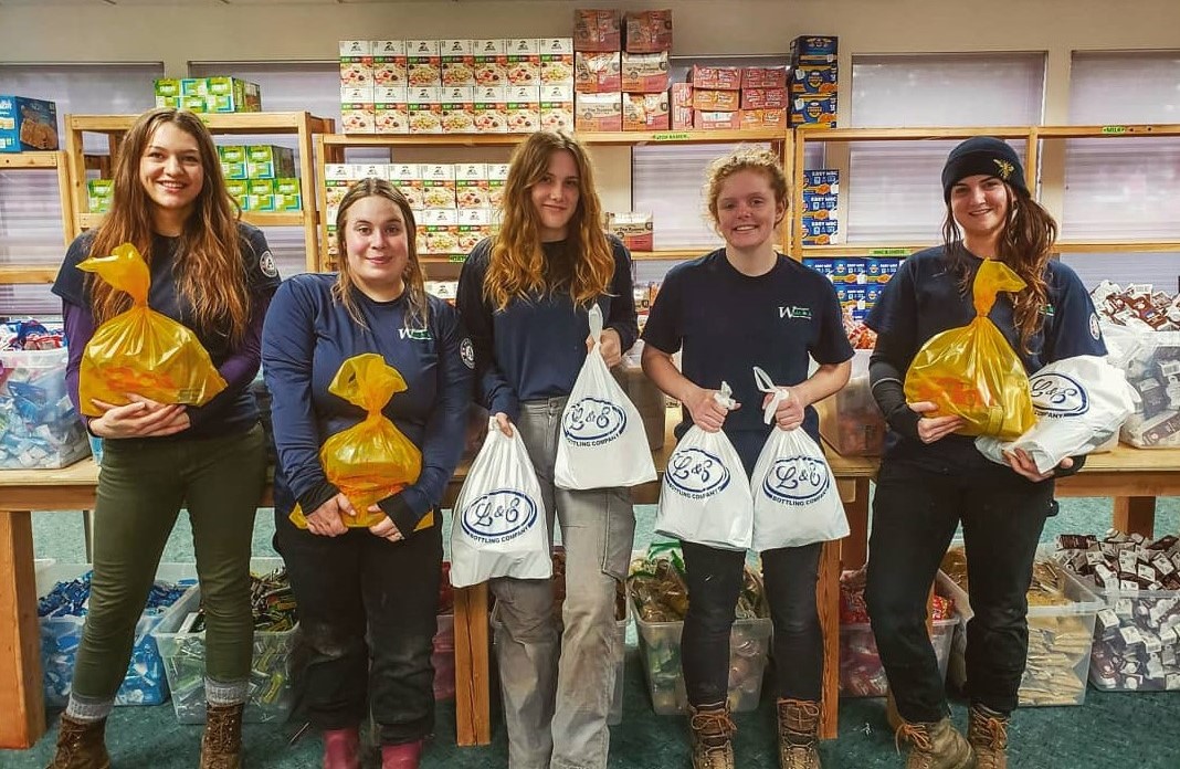 Five young adults stand in a line wearing dark blue shirts and carrying yellow and white plastic bags full of food.