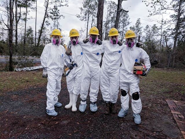  Five WCC members pause for a group photo in a yard outdoors, wearing white Tyvek suits during a disaster response project
