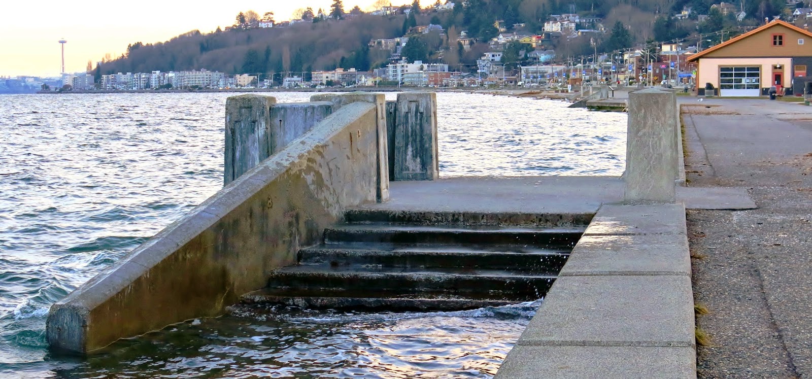 Water coming up high on cement steps at Alki Beach with waterfront buildings and Spaceneedle showing in the background.
