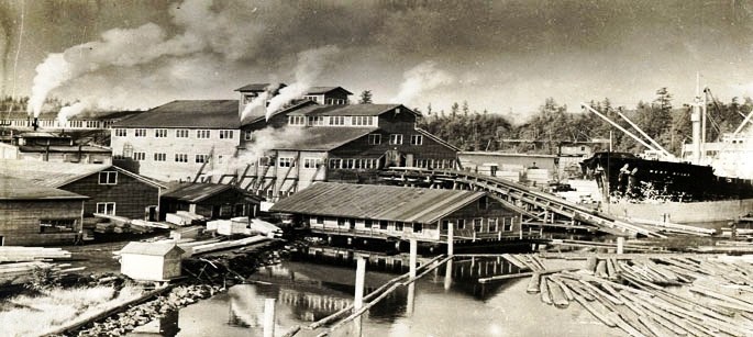 Black and white photo of mill with logs in foreground and building with smoke billowing in background.