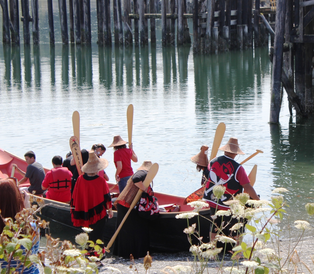 Large canoe on water with nine tribe members holding oars, wearing black and red.