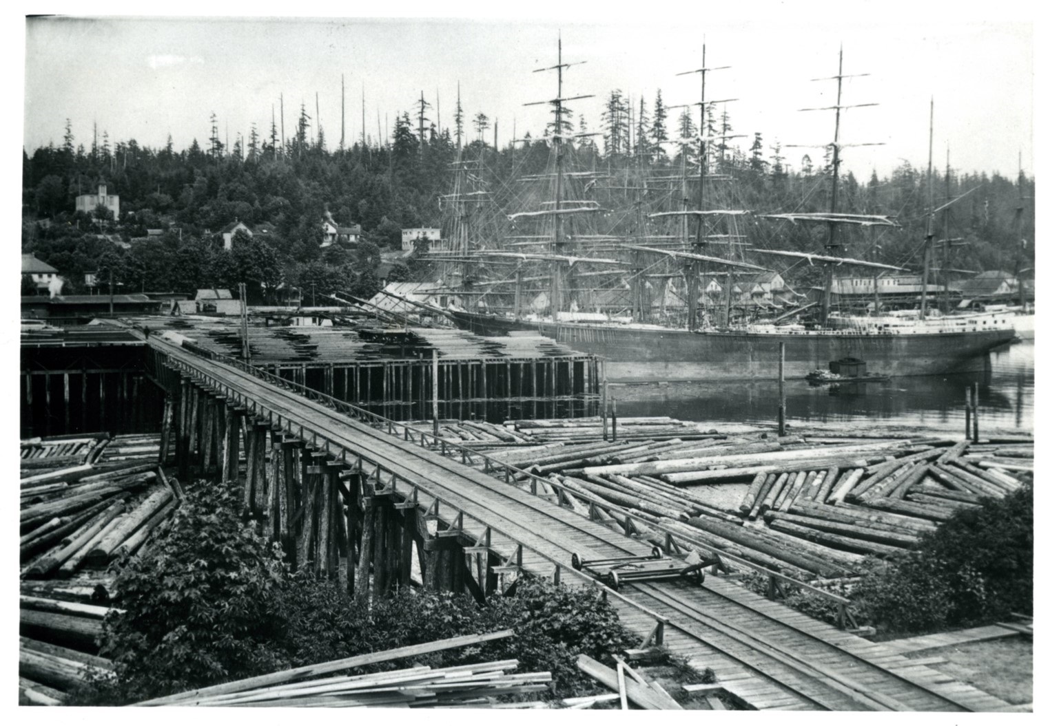 A black and white image of a port with a train trestle over the water, logs in the water on either side, and a large three-masted boat. 