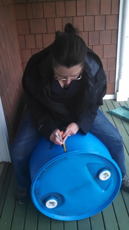 person sitting on a rain barrel laying on its side, measuring it. 