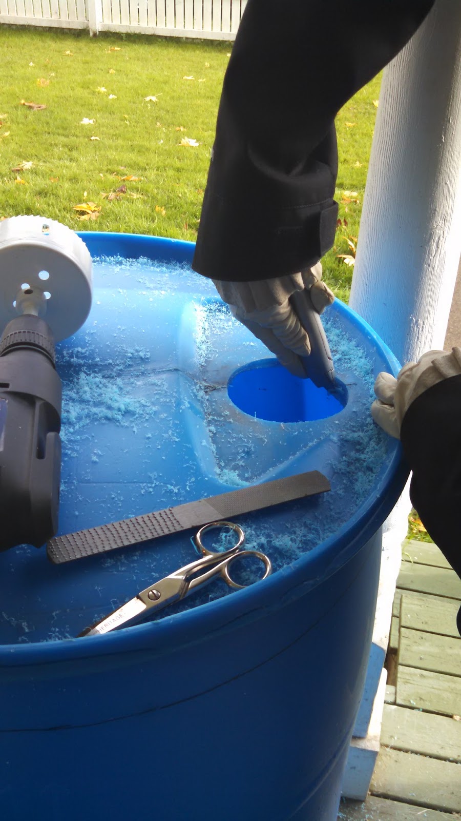 A hand cuts a circle out of the top of a blue rain barrel. 
