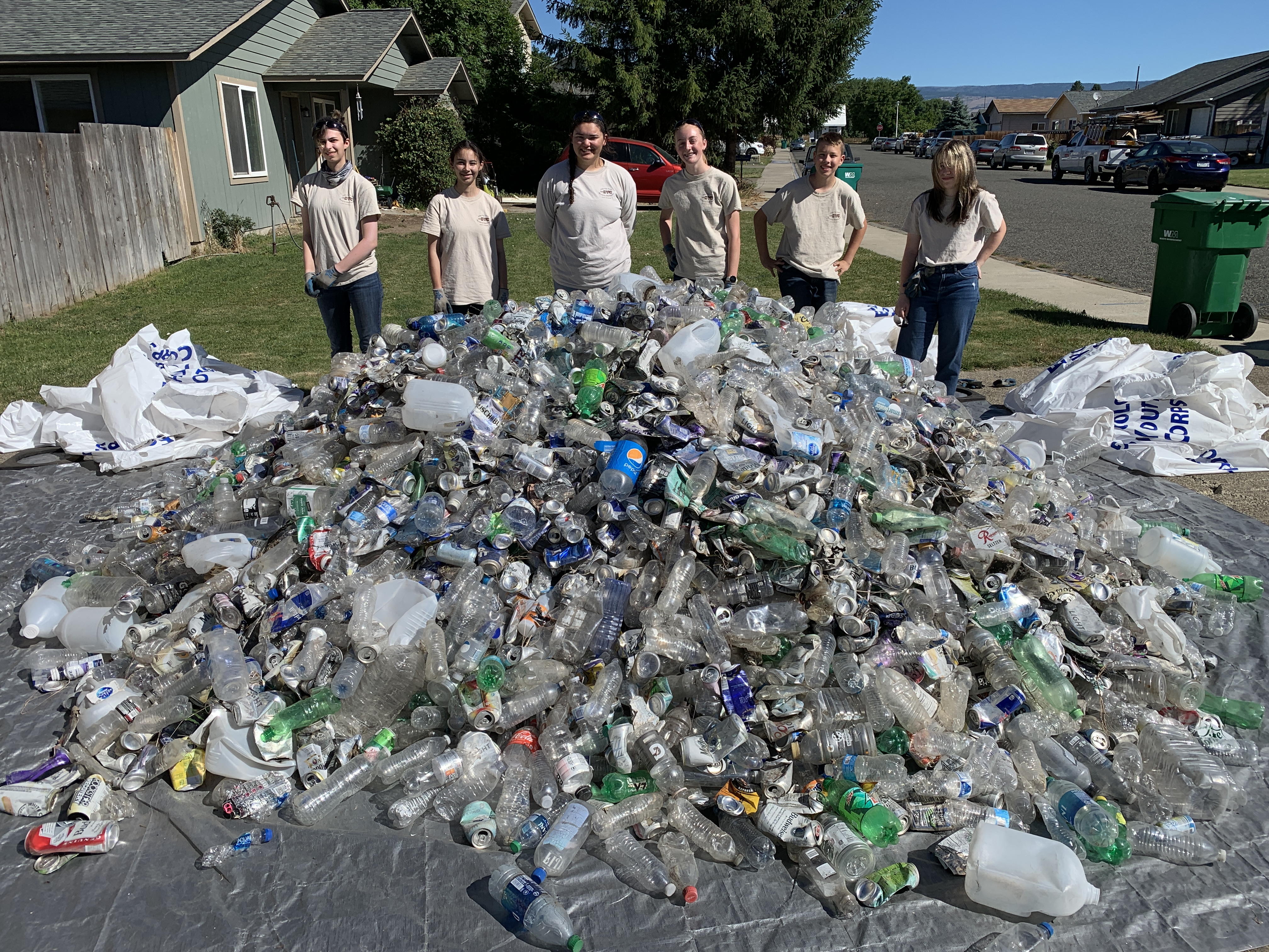 On a sunny day, six teens in light shirts smile at the camera from behind a large pile of plastic bottles and aluminum cans spread on a gray tarp.