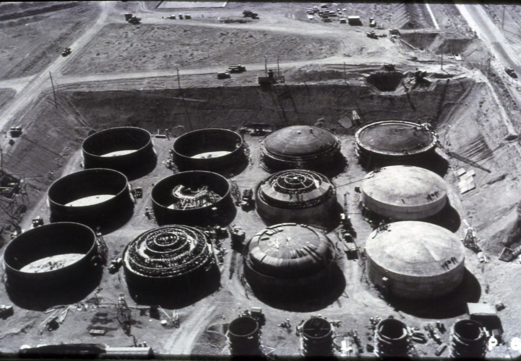 Black and white aerial view of the B Tank Farm at Hanford under construction in the 1940s, showing massive storage tanks in varying states of construction. 