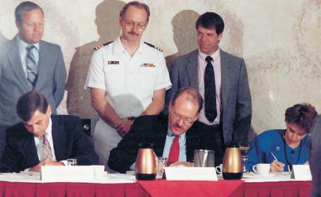 Three people sit at a table signing documents, three stand behind watching