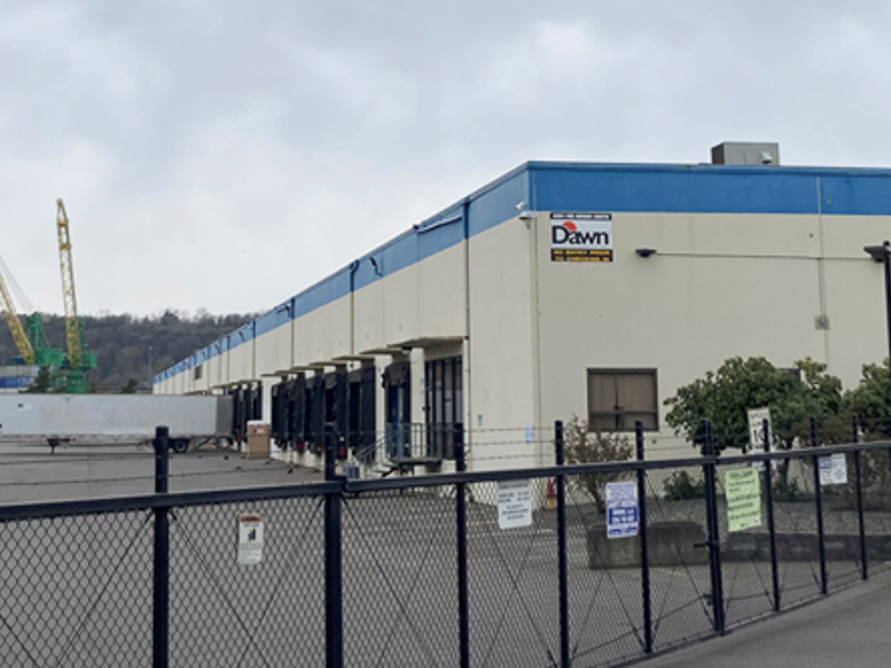 Industrial warehouse buildings behind chain link fence with cranes in background, next to Lower Duwamish Waterway