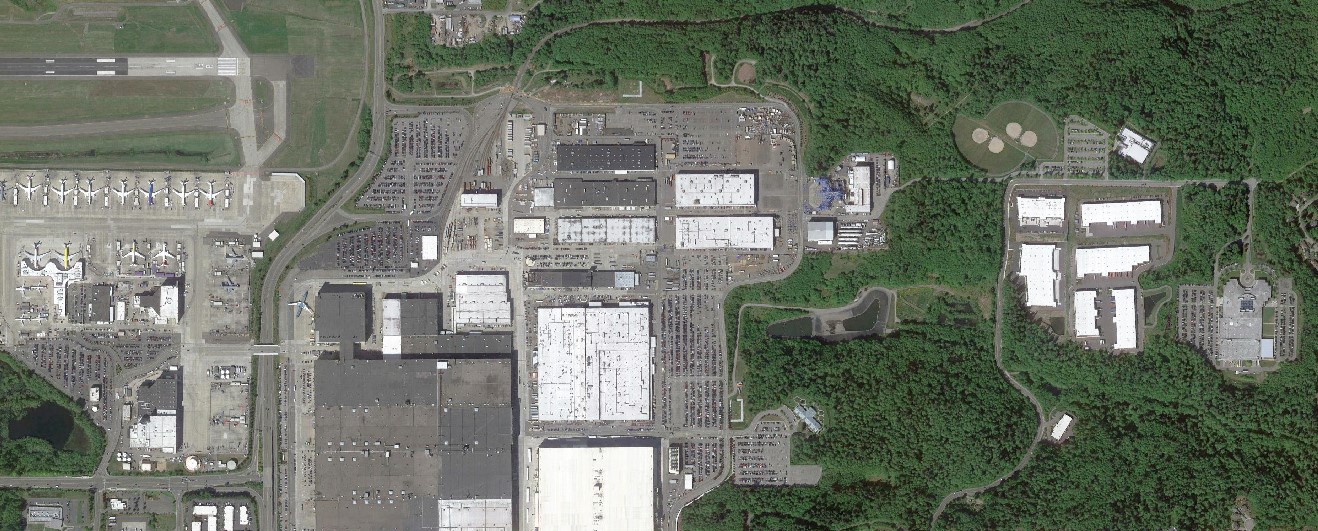 Aerial view. Industrial buildings in center. Jetliners parked on left. Forested area on right. 