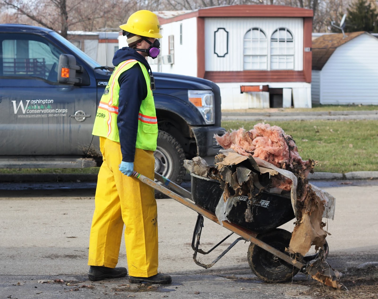 A WCC AmeriCorps member carries a wheelbarrow full of debris. They are wearing a yellow hard hat, respirator mask, and yellow pants & vest.