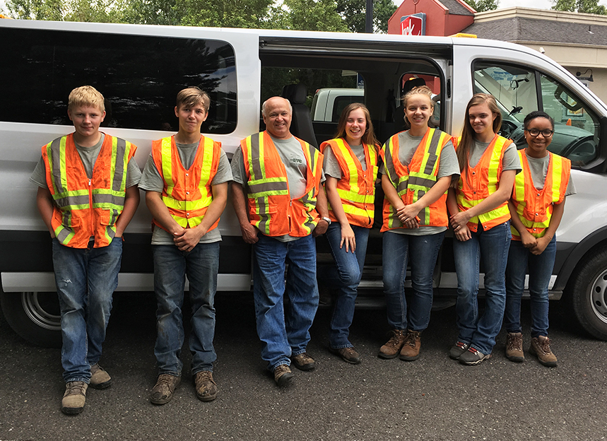 An Ecology Youth Corps youth crew consisting of six teen crew members dressed in boots, jeans and blaze-orange vests pose for a photo with their crew leader.