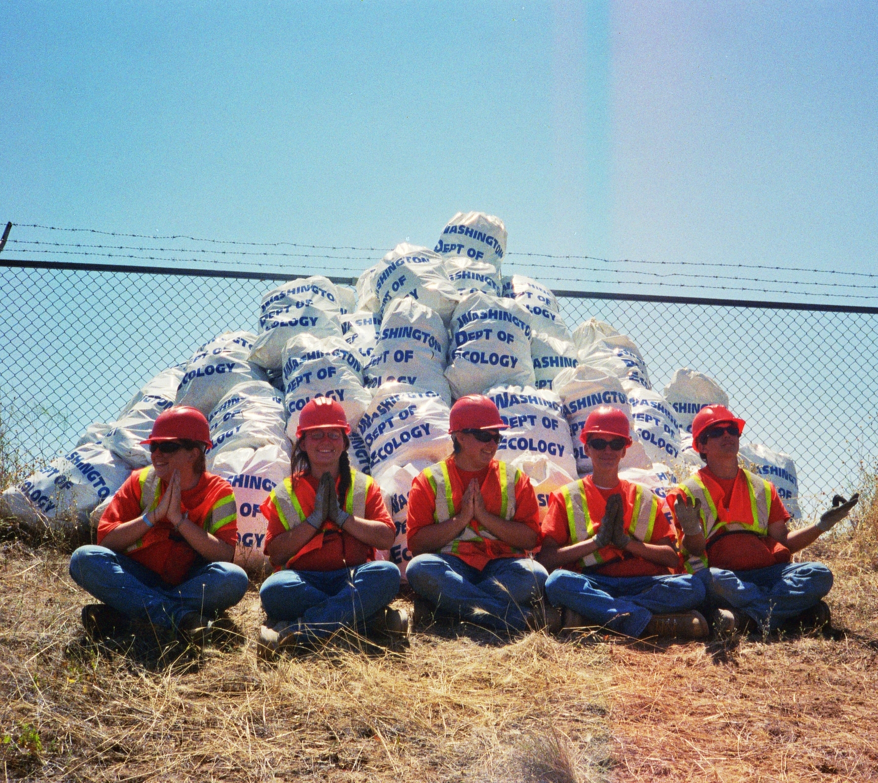 Five youth sit cross-legged in front of a pyramid pile of stacked litter bags. The youth are wearing hardhats, orange vests, jeans and work boots.