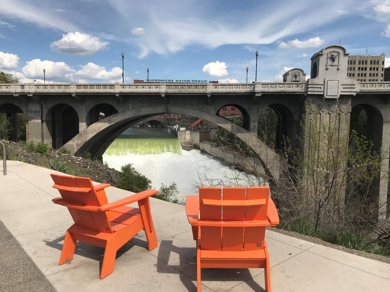 Photo of a the Spokane River with a bridge in the background and two orange chairs in the foreground. 