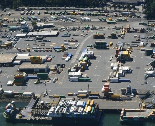 Aerial view of a large industrial paved area, with many shipping containers seens. Barges with containers are moored to a pier at the bottom. In the left center, some one storey warehouses are intersp