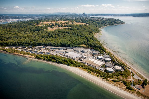 Aerial view of West Point Wastewater Treatment Plant on a narrow strip of land with Puget Sound surrounding, forest and Seattle in back