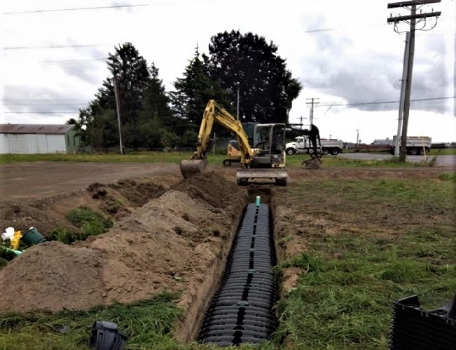 Drain field pipe being installed in yard, with excavator in the background