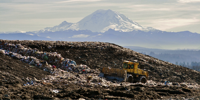 An open pit of waste in a landfill, a bulldozer is moving the waste. Mt. Rainier is in the background.