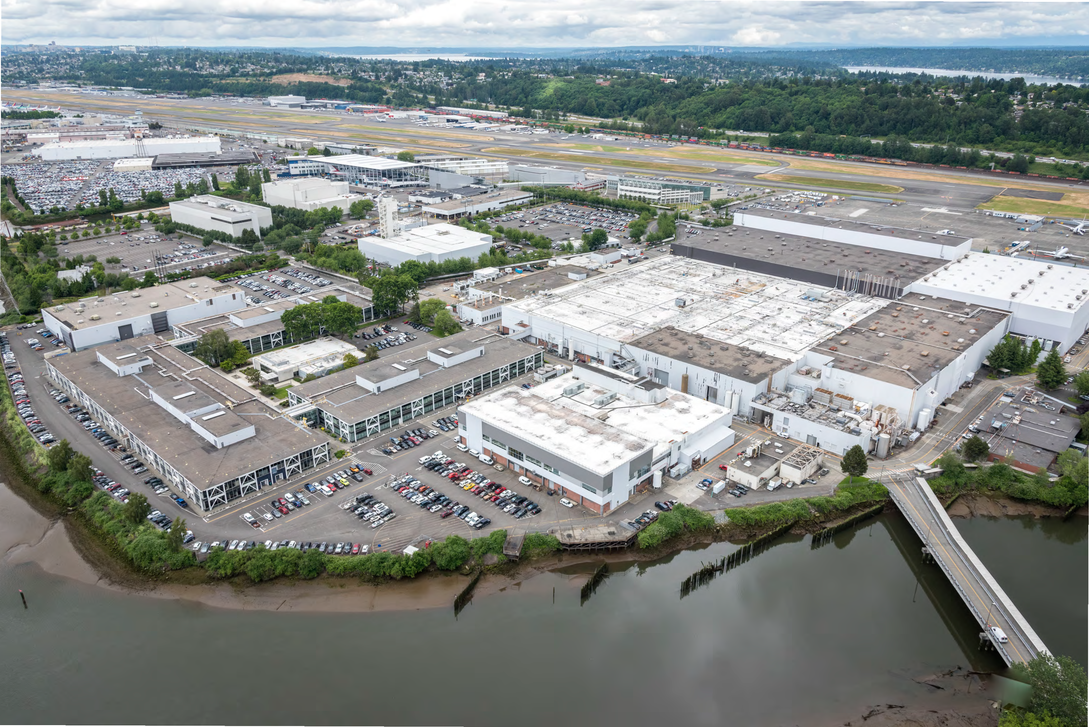 A commercial and industrial site with many buildings and parking areas. A river is seen in the foreground, and the background shows more industial buildings, an airfield and a wooded ridge with homes.