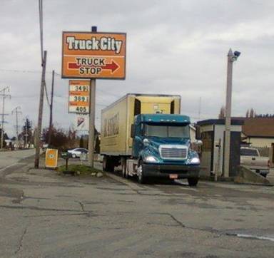 Semi truck, pulled up to fuel dispensers, under a sign reading Truck City Truck Stop. 