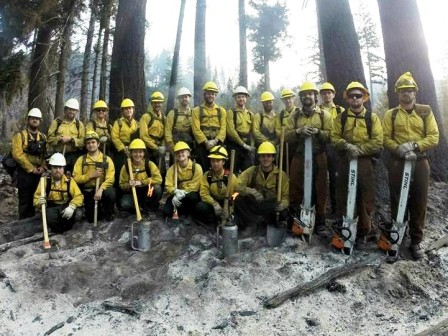 A crew of 20 WCC AmeriCorps members gather for a photo holding shovels, axes and chainsaws in a burned out area during the Stewart Mountain wildland fire response.