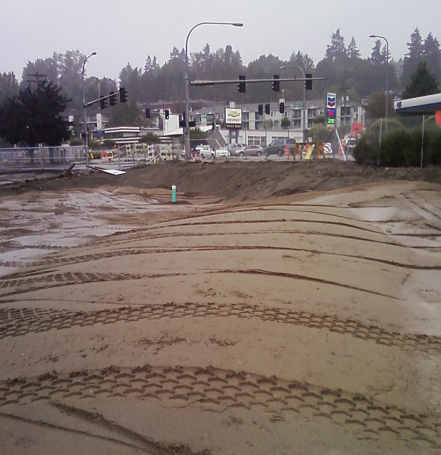 Clean dirt backfilled over the site excavation. Intersection in background.