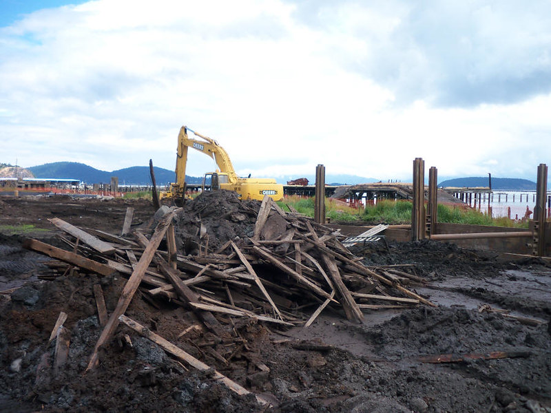 Heavy machinery behind a pile of wood waste in mud on the shore of Puget Sound.