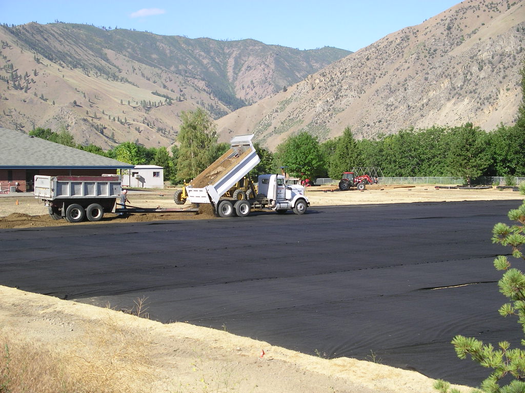 A dump truck places dirt over a large space covered in black fabric.
