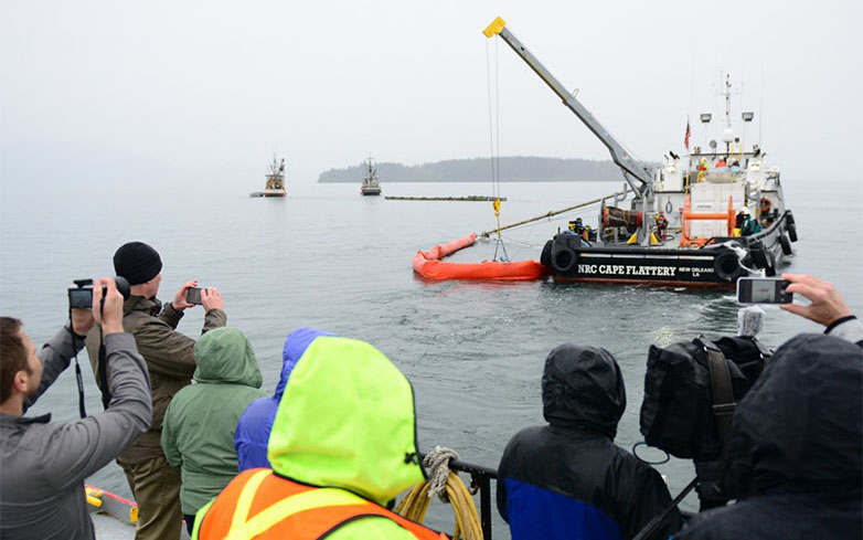 Several people with cameras video recording a vessel deploying boom in water.