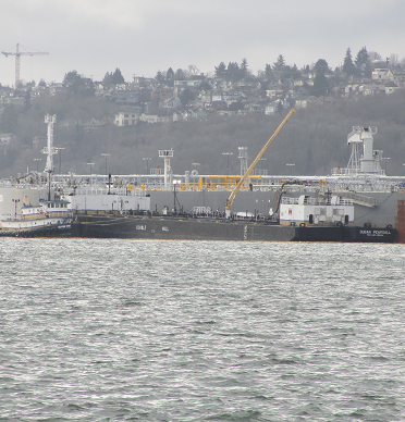 Tanker loading cargo in Seattle. Tank vessel with a tug and barge tied up alongside. The Seattle shoreline is in the background and the waters of the Salish Sea are in the foreground.