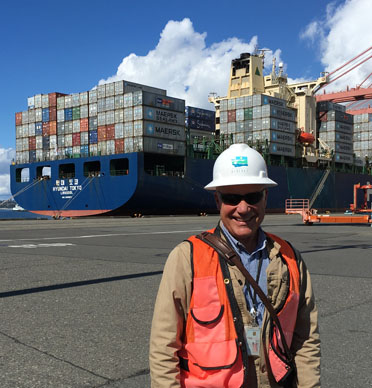Man in a hard hat and safety vest stands in front of a container ship.