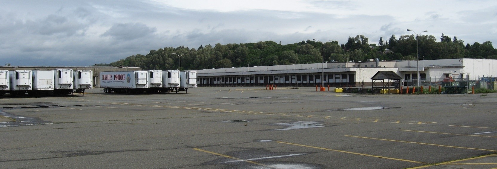 Several refrigerated semi truck trailers are parked in a row on the left. On the right is a long loading dock of a warehouse. A tree covered hill is in the background.