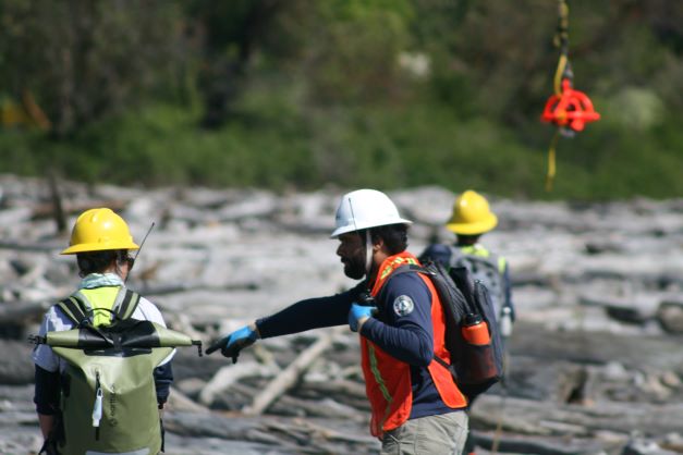 A WCC crew supervisor wearing a white hard hat points at the ground. They are directing their crew to debris that needs to be removed from the beach.