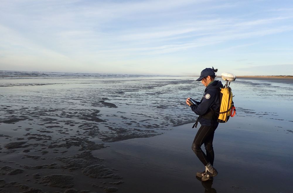 A WCC member stands on a sandy beach along the Pacific Ocean. They are facing out toward the water while carrying monitoring equipment in their hands and in their backpack.