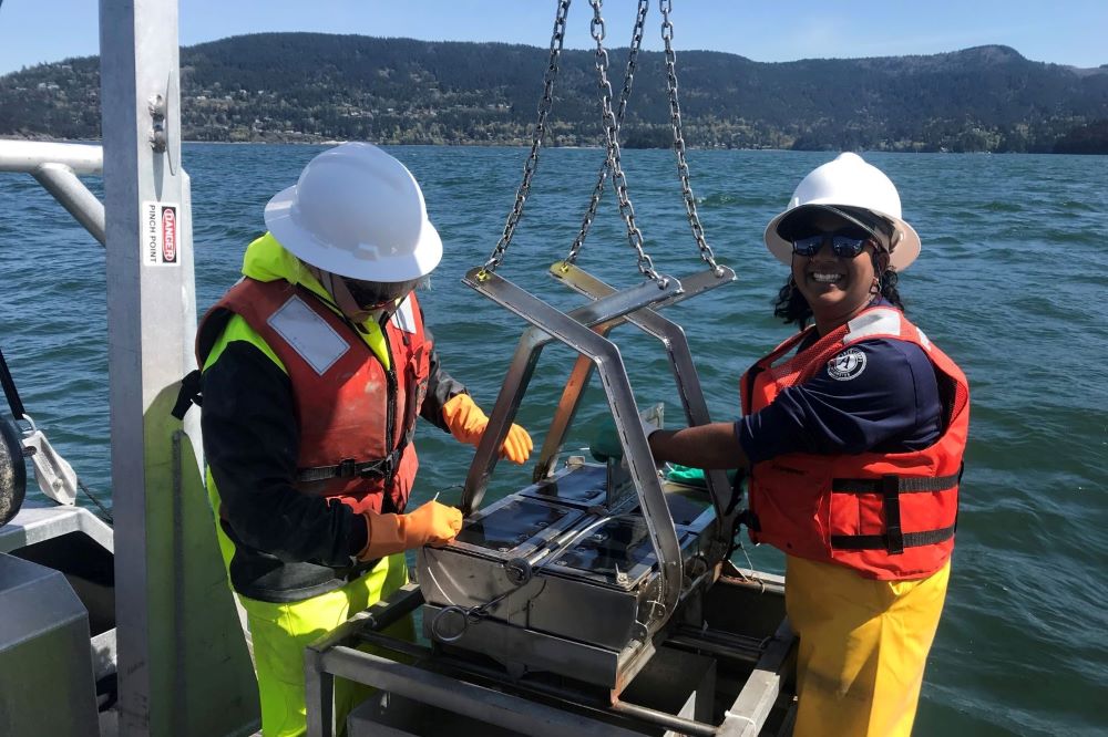 Two WCC members wearing life vests, gloves, and hard hats stand on the edge of a boat in the Puget Sound. They are collecting sediment samples using a special tool attached to the boat.
