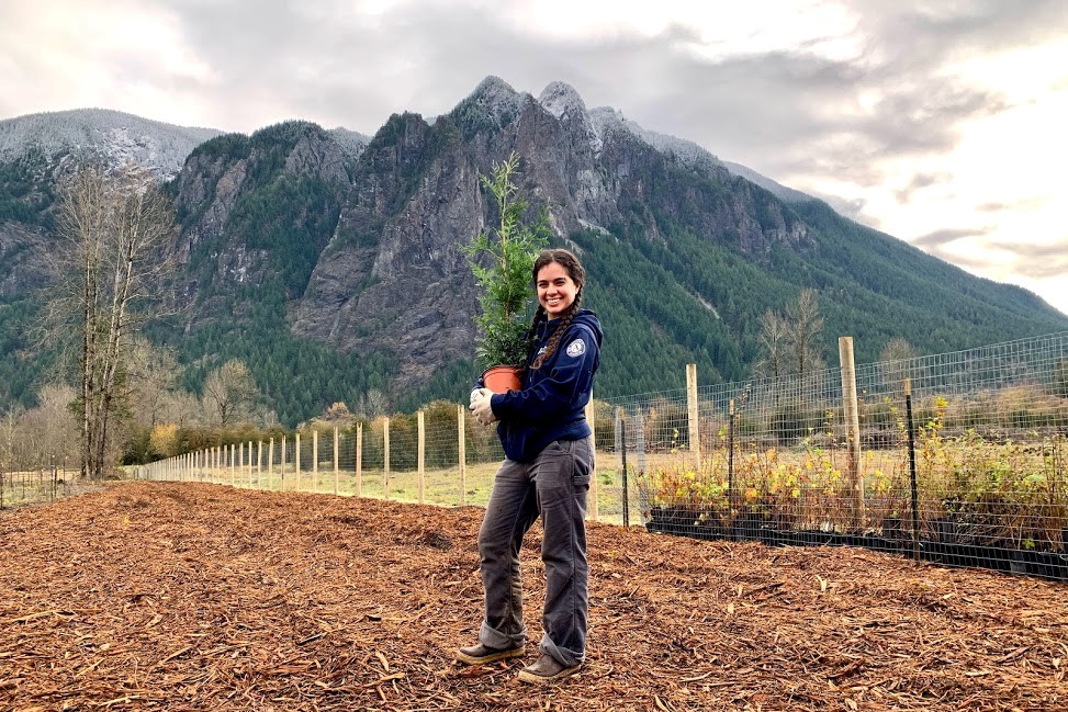 A WCC members hold a potted plant while standing in an open field. Behind them is a snow-capped rocky peak.