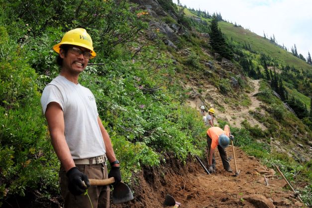 A WCC member in a hard hat, safety glasses, and gloves smiles, holding a grub hoe and standing on a dirt trail through a meadow on a steep slope. 