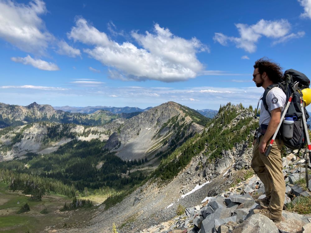 A WCC member stands on a rocky pass trail looking down into a valley filled with trees. They are wearing a large pack with a hard hat and a pair of loppers strapped to the exterior.