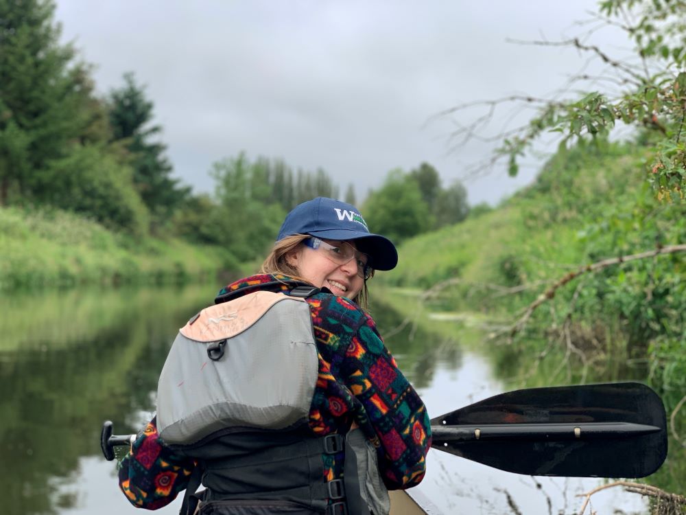 A WCC member sits in the front of a canoe on a small body of water surrounded by greenery. They are facing away from the camera but turning around to smile. They are wearing a small backpack, safety glasses, and a WCC cap and are holding a paddle.